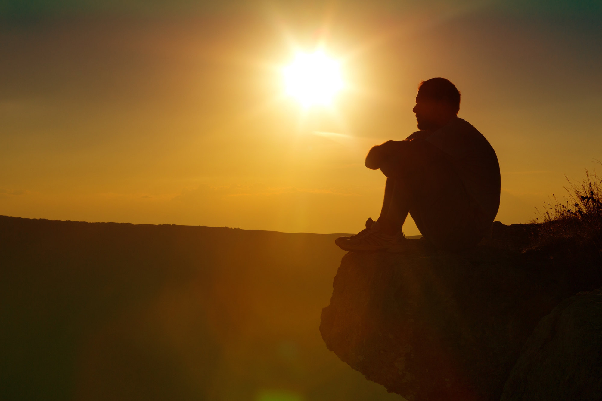 A man on a mountain rock, reflecting on his Christian lifestyle as the sun sets.